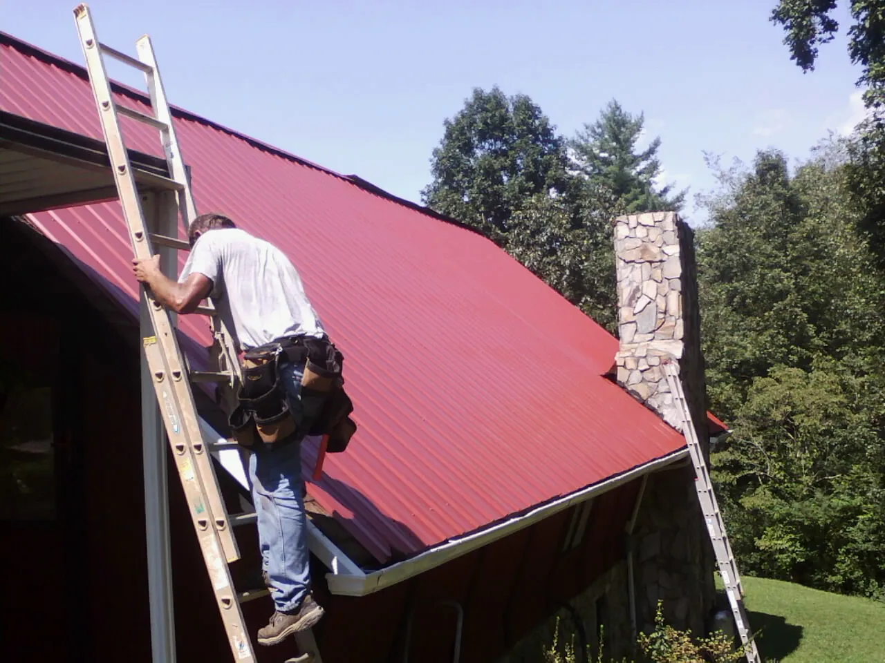 Roofer on a ladder beside a red metal roof and stone chimney — Soffit & Fascia Repair by Roofing Brothers in La Mirada